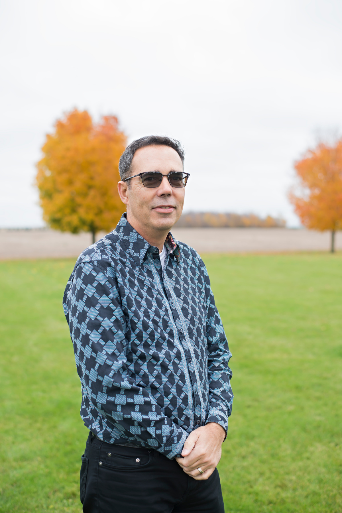 Man with dark glasses and blue plaid shirt with colourful tree in background